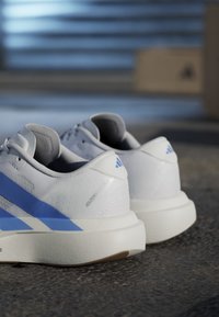 Scarpe da corsa Adidas bianche e blu posizionate su terreno testurizzato con uno sfondo sfocato di una scatola e linee orizzontali.