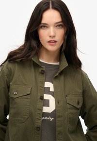 Camicia verde oliva con bottoni, dotata di due tasche frontali e bottoni marroni, indossata sopra un felpa grigia con un design numerale bianco.