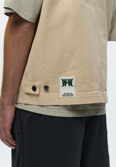 Beżowa koszula z krótkim rękawem z czarnymi guzikami i białą metką z napisem "Wood Wood Copenhagen, 2002" noszona z czarnymi spodniami.