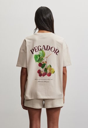 Femme aux longs cheveux foncés portant un short beige et un t-shirt beige avec l'inscription « PEGADOR » et un motif de branche de framboisier dans le dos.
