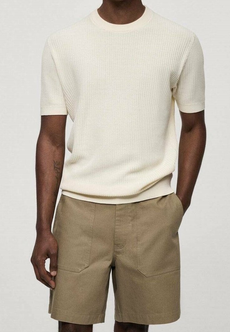 Homme portant un pull en tricot crème à manches courtes et un short kaki longueur genou, debout avec une main dans la poche et l'autre détendue.