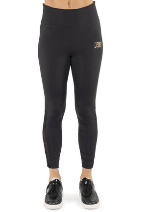 Modèle portant un legging noir taille haute orné d’un petit logo doré et des baskets noires sur fond blanc.