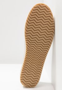 Semelle de chaussure en caoutchouc avec un motif ondulé texturé, couleur beige. Le bord est légèrement arrondi, avec un matériau supérieur de couleur claire visible sur les côtés.