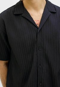 Uomo che indossa una camicia nera a maniche corte con texture e colletto, che rivela parzialmente un tatuaggio rosso sul petto.