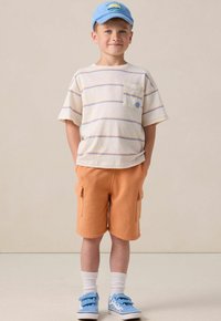 T-shirt à rayures à manches courtes avec poche, shorts cargo orange, casquette bleue, chaussettes blanches, baskets bleu à velcro avec détails en motif.