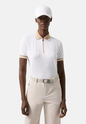 Vrouw gekleed in een wit en beige golfoutfit, inclusief een witte pet, een poloshirt met rits, beige broek en een geweven riem, staand tegen een effen achtergrond.