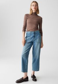 Donna che indossa un maglione a collo alto marrone, jeans blu a vita alta e larghi con grandi tasche frontali, e mocassini in pelle marrone scuro, in piedi su uno sfondo grigio chiaro.