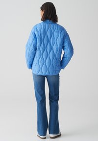 Veste matelassée bleu clair avec un motif texturé, dotée d'un col montant et de manches longues, assortie à un jean bleu évasé et des chaussures blanches.