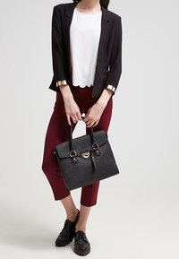 Sac à main noir texturé avec une forme structurée, doubles poignées et quincaillerie dorée. Porté avec un haut blanc, un blazer noir et un pantalon bordeaux.
