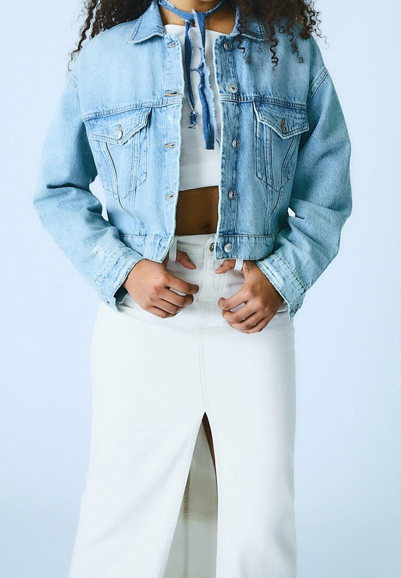 Donna che indossa una giacca di jeans cropped blu chiaro sopra una maglietta cropped bianca e una gonna bianca a vita alta con uno spacco frontale, in piedi contro uno sfondo chiaro.