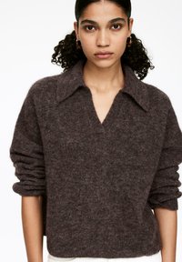 Frau mit zurückgehaltenem lockigem Haar, die Schildpatt-Ohrstecker in Form von Ohrringen und einen braunen, fuzzy Strickpullover mit weitem Kragen und V-Ausschnitt trägt.