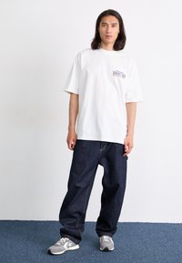 Hvit oversized t-skjorte med et lite blått og rødt grafisk motiv på brystet, sammen med mørkeblå denimjeans og grå joggesko.