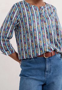 Blusa a righe blu e bianche con motivo floreale, scollatura rotonda, dettagli con bottoni, maniche a tre quarti e vestibilità rilassata. Indossata con jeans a vita alta.