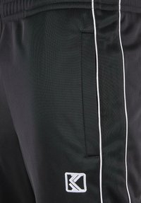 Pantaloni de trening negri dintr-un material texturat, cu dungi albe pe laterale și un patch cu logo pe partea stângă. Include un buzunar lateral.