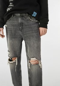 Vyprané černé džínové kalhoty s opotřebovanými koleny, s roztřepenými okraji a dvěma velkými trhlinami. Nošeny s černou mikinou s viditelnou etiketou.