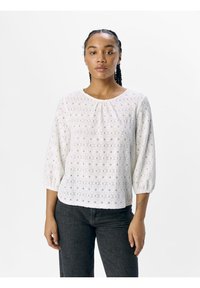Blusa bianca in pizzo con scollo arricciato, maniche a tre quarti a palloncino e motivo floreale intagliato. Abbinata a pantaloni grigio scuro.
