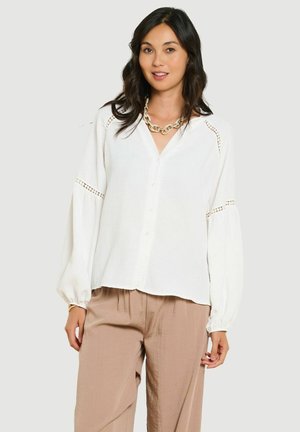 Vrouw draagt een witte blouse met lange mouwen en kanten details en beige losse broek, poserend tegen een lichtgrijze achtergrond.