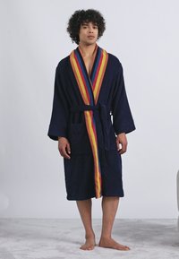 Noor inimene seisab paljajalu, seljas tume sinine hommikumantel, millel on mitmevärviline triibuline äär ja vöö, ühtlase tausta ees.