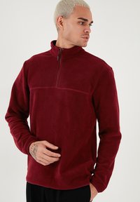 Pullover in pile color bordeaux con zip metà, maniche lunghe, texture liscia e una cucitura orizzontale sul petto.