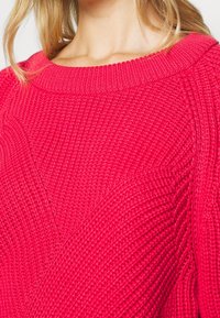 Gros plan sur une personne portant un pull tricoté rouge vif texturé avec un large col rond, sur un fond uni.