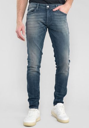 Jean en denim bleu foncé ajusté avec une légère décoloration, design à cinq poches et fermeture éclair. Porté avec des baskets blanches.