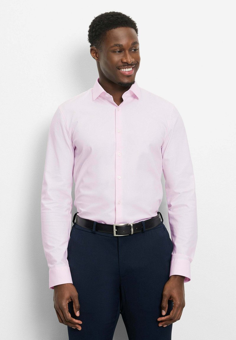 Homme souriant, portant une chemise à manches longues rose clair avec des boutons, rentrée dans un pantalon bleu marine foncé avec une ceinture noire, debout contre un mur blanc.