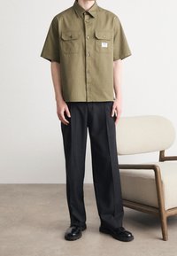 Olivenfarbendes Kurzarmhemd mit zwei Brusttaschen und Knopfverschluss, kombiniert mit schwarzen weiten Hosen und schwarzen Schuhen. Minimalistisches Design.