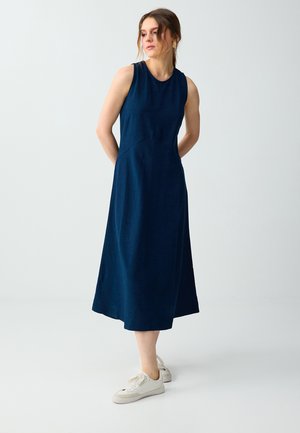 Jeune femme debout, les mains dans le dos, portant une robe midi bleu marine sans manches et des baskets blanches sur un fond uni.