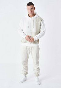 Sudadera blanca con acentos beiges, cordones y bolsillo frontal; combinada con pantalones jogger color crema y zapatillas blancas; textura de tela suave.