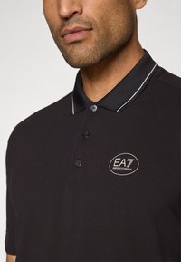 Melna polo krekls ar adītu tekstūru, trīs pogu aizdari un baltiem svītrotiem apkakles maliem. Uz krūtīm redzams EA7 logo.