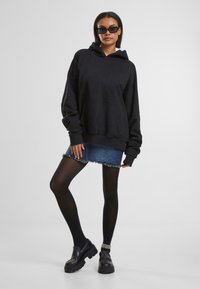 Zwarte oversized hoodie met een voorzak, gecombineerd met een denim minirok. Het model draagt zwarte panty’s en zwarte platform schoenen, met een zonnebril.