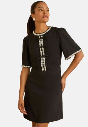 Schwarzes Kleid mit kurzen Puffärmeln, verziertem Ausschnitt, dekorativen Knöpfen und kontrastierendem Blumenbesatz an Ärmeln und Vorderseite.