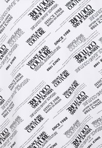 Vitt tyg med upprepande svart text som säger "VERSACE JEANS COUTURE," "SINCE 1988," och "BASED IN MILAN," i olika typsnitt och riktningar.