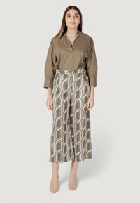 Camicia verde oliva con bottoni e taschino frontale, abbinata a pantaloni larghi a fantasia in olive e crema. Tessuto liscio, décolleté neutri.