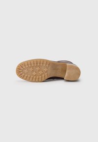 Chaussure en cuir marron foncé avec une semelle en caoutchouc texturé, présentant un motif de bande de roulement marron clair. Nom de la marque estampé visible.