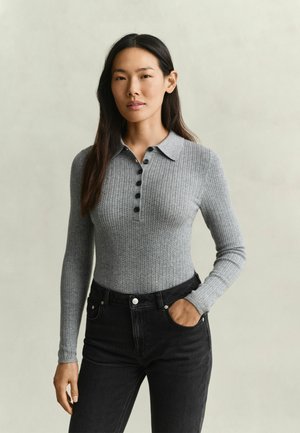 Frau mit langen dunklen Haaren, trägt einen grauen, gerippten Langarm-Polopullover und schwarze hoch taillierte Jeans, eine Hand in der Tasche, neutraler Hintergrund.