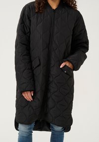 Černý prošívaný dlouhý kabát se zipem vpředu, žebrovaným límcem a bočními kapsami. Má měkký strukturovaný povrch a zakřivený spodní lem.