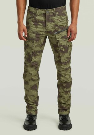 Mann trägt grüne und braune Tarn-Cargohosen mit mehreren Taschen und schwarze Schnürstiefel vor einfarbigem Hintergrund.