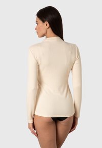 Maglietta a maniche lunghe a coste di colore beige, con collo alto e silhouette aderente, abbinata a un pantalone nero. Texture liscia e design minimale.