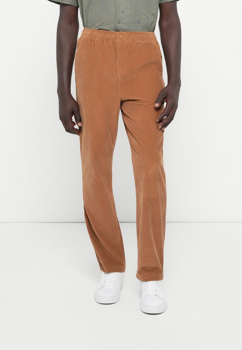 Homme portant un pantalon en velours côtelé marron avec une ceinture élastique et des baskets blanches sur fond blanc.