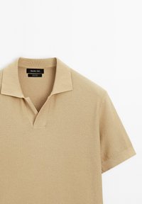 Polo beige realizzato in 100% cotone con un motivo testurizzato, maniche corte e colletto a V senza bottoni. Etichetta visibile all'interno.