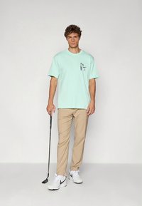 T-shirt de algodão verde claro com um pequeno gráfico, combinada com calças bege. O modelo segura um taco de golfe, calçando ténis brancos desportivos.