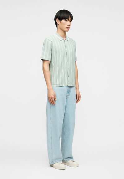 Νέος άνδρας που φοράει ανοιχτό πράσινο πουκάμισο με κοντά μανίκια και λευκές ρίγες, ανοιχτό μπλε τζιν παντελόνι με φαρδιά μπάτσα και λευκά αθλητικά παπούτσια.