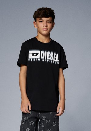 T-shirt nera con logo bianco "DIESEL", maniche corte, scollatura rotonda. Indossata con jeans scuri decorati con un motivo di faccine sorridenti.