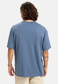 Person med krøllet brunt hår iført en løs blå t-shirt og beige bukser, stående med ryggen mod seeren på en ensfarvet baggrund.