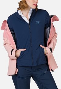 Marineblaues Jacket mit rosa Akzenten, aus einem glatten Stoff gefertigt. Es verfügt über einen Reißverschluss, einen hohen Kragen und Reißverschlusstaschen an der Hose.