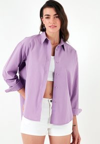 Light purple button-up shirt with a collar, relaxed fit, long sleeves rolled up, and lightweight fabric. Worn over a white crop top and shorts.