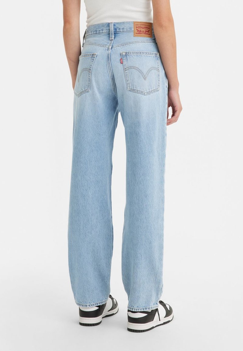 Hellblaue Denim-Jeans mit lockerer Passform, hoher Taille, zwei Gesäßtaschen und einem Lederlogo-Patch am Bund. Schwarz-weiße Turnschuhe.