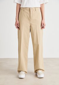 Pantalon large beige en coton, doté d'une fermeture à bouton, de poches avant et d'une coupe décontractée. Associé à des sneakers blanches.