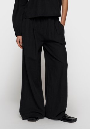 Osoba nosząca luźne czarne spodnie z szerokimi nogawkami i bocznymi kieszeniami, w połączeniu z czarnymi butami o kwadratowym nosku, stoi na jasnoszarym tle.